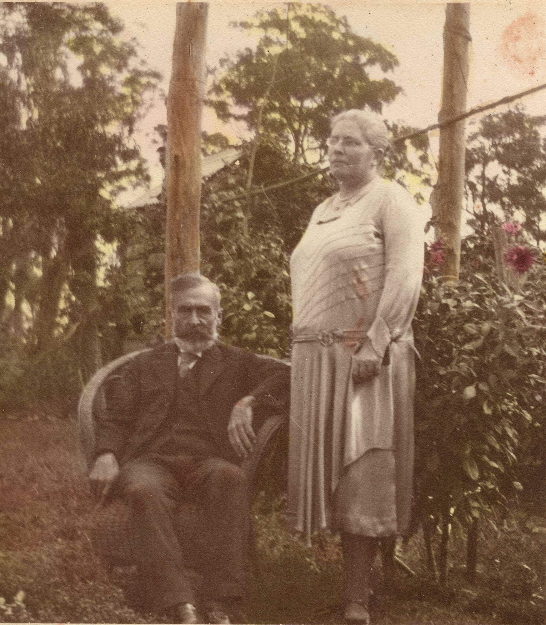 Hand-tinted photograph of an elderly couple in a garden: a white-bearded man seated in a cane chair, a woman standing beside him in a pale dress, dahlias behind them.