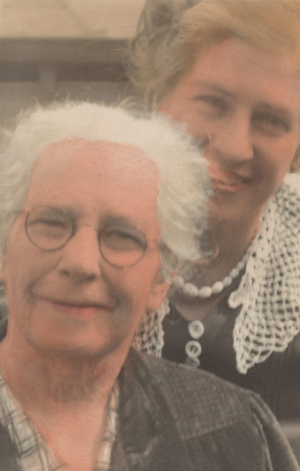 Hand-tinted close-up of an elderly white-haired woman in round glasses and a dark dress with a check collar, with a younger fair-haired woman in a dark dress and a deep crocheted lace collar behind her.