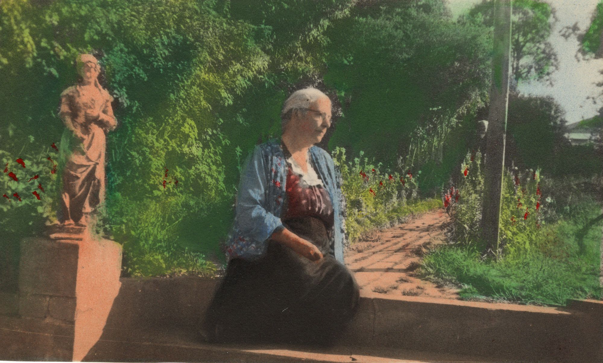Hand-tinted photograph of an elderly woman seated on a low brick wall in a garden, a classical statue on a plinth behind her and hollyhocks along a brick path.