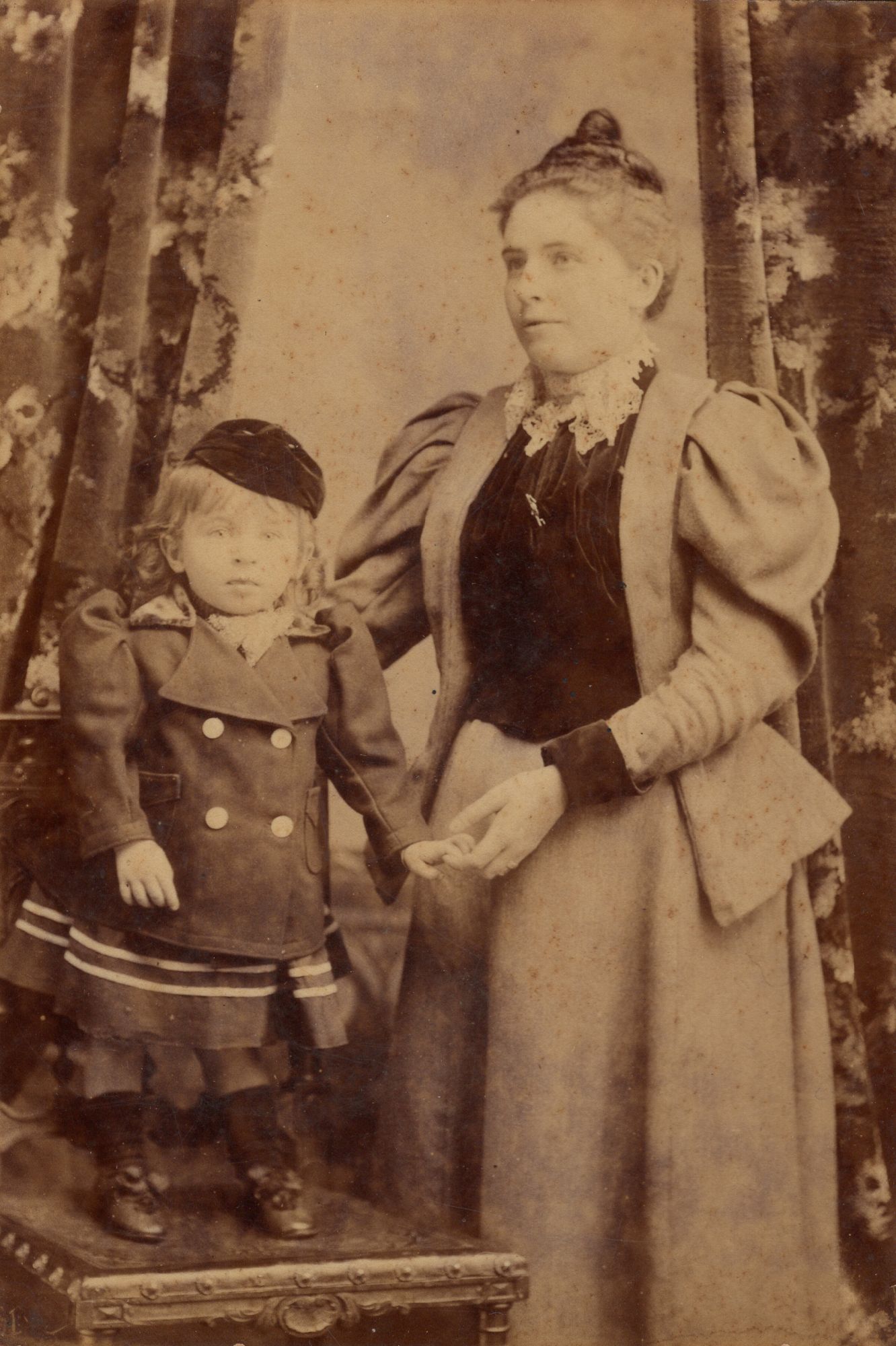 Sepia studio portrait: a young woman in a jacket with very full leg-of-mutton sleeves, and a small child in a double-breasted coat and cap standing on a chair beside her.