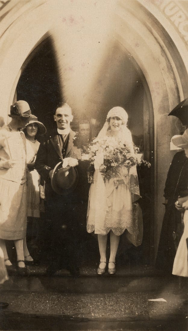 A minister in a dark suit and clerical collar, holding a bowler hat, stands beside his bride in an ivory knee-length dress and a long lace veil in the pointed-arch doorway of a stone church, wedding guests to either side.