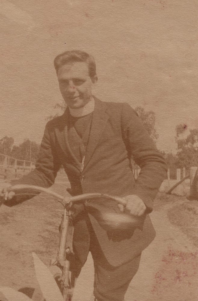 A young man in a suit and clerical collar stands smiling behind a bicycle in an open paddock, one hand on the handlebar, scattered gum trees and a fence line behind him.