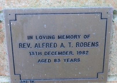 A bronze memorial plaque on a brick wall, a small cross above the text: “In loving memory of Rev. Alfred A. T. Robens, 13th December 1982, aged 83 years.”