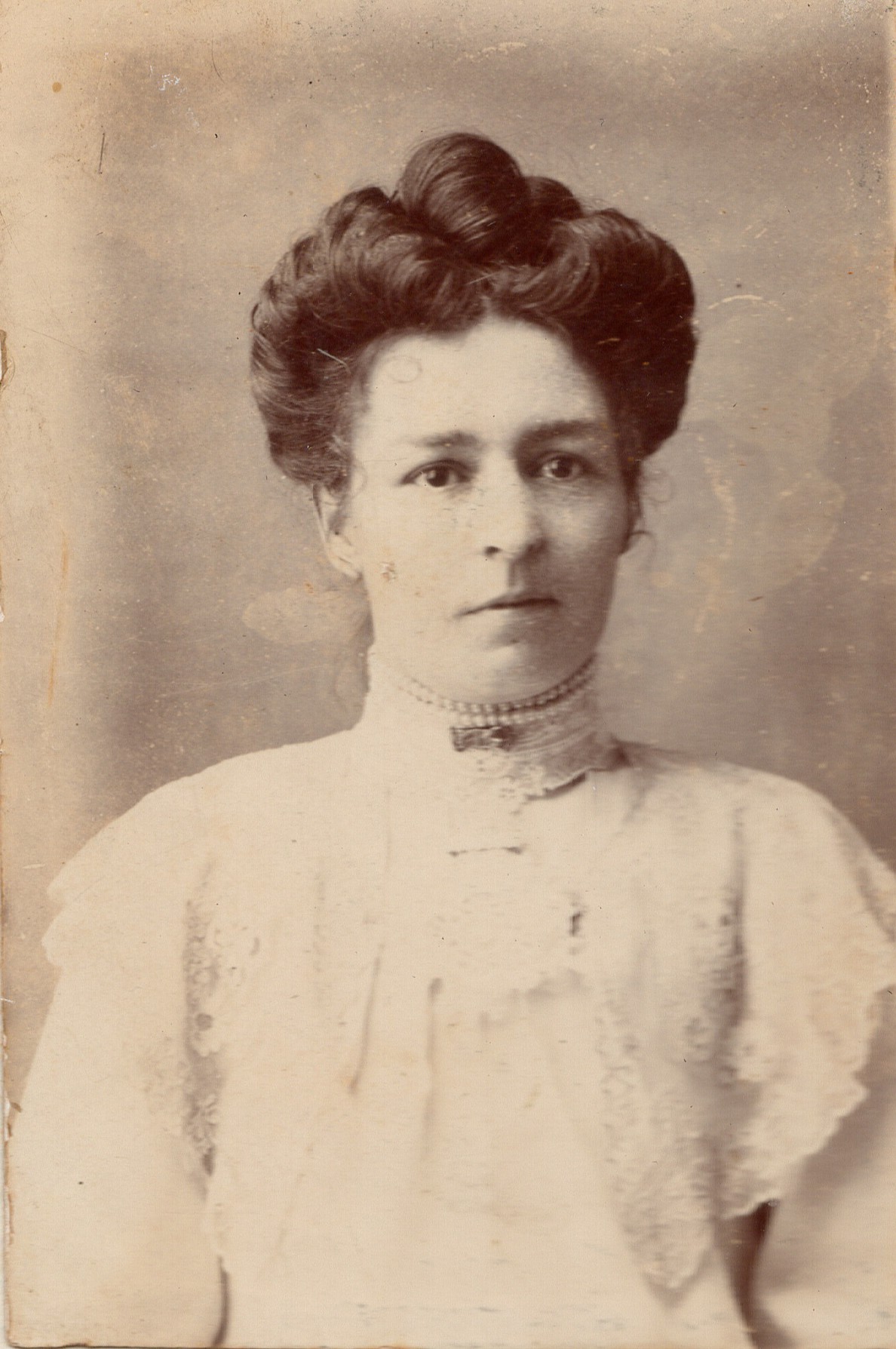 Studio half-length portrait of a young woman with a full pompadour, wearing a high-necked blouse with a deep lace yoke and a beaded choker.