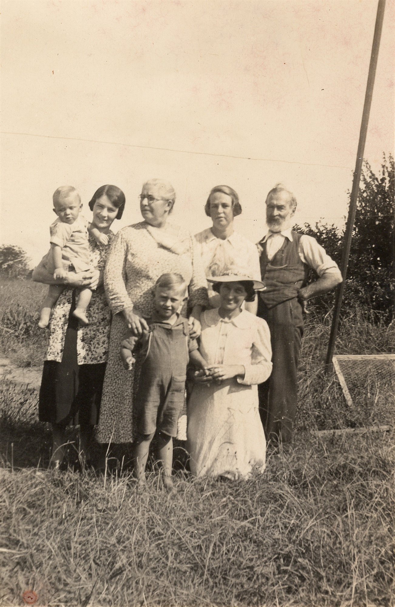 Six people stand and kneel in a field: a woman holding a toddler, an older woman, another woman, a bearded elderly man, a young boy, and a woman in a sun hat kneeling in front.