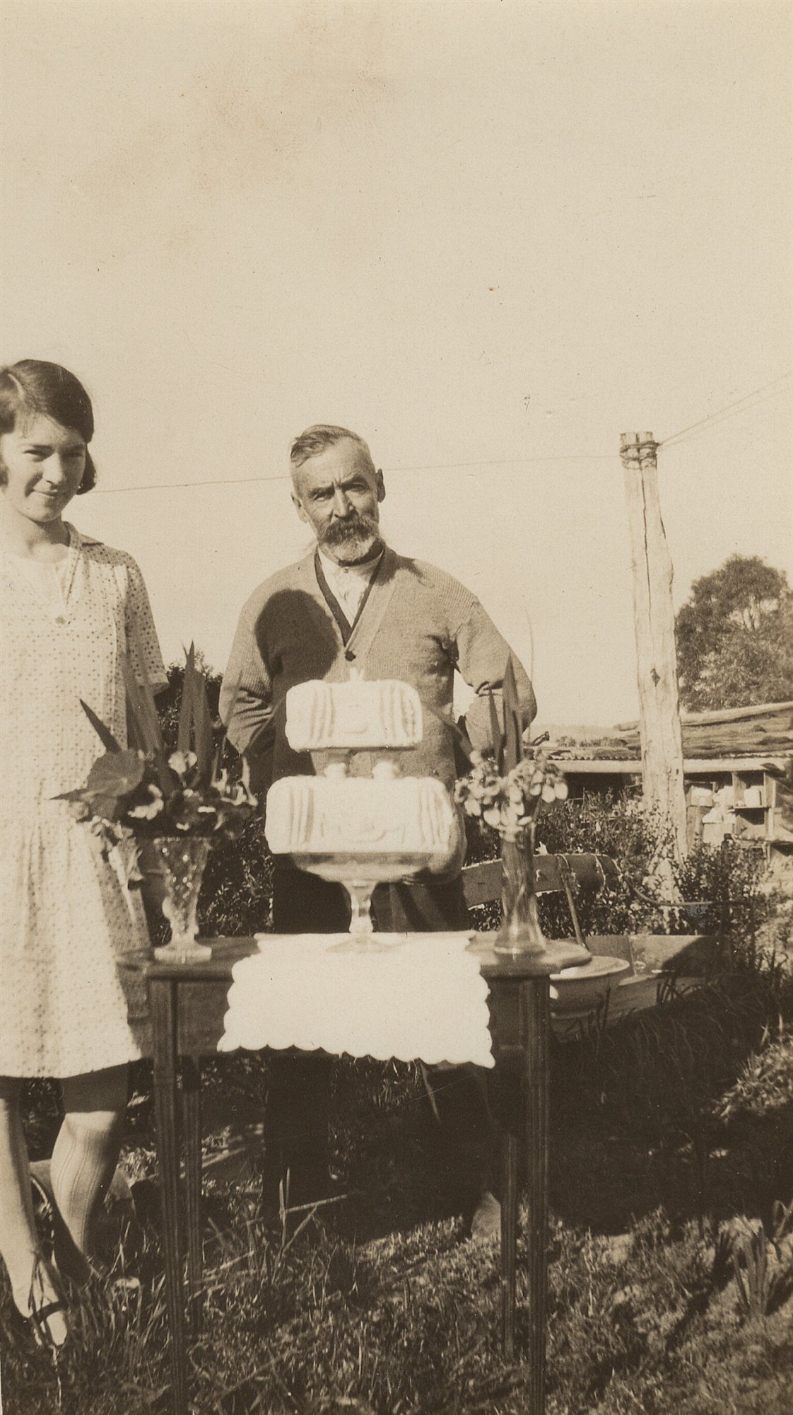 A bearded elderly man and a young woman stand behind a garden table set with a two-tiered decorated cake and vases of flowers.