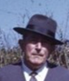 A colour photograph of an elderly man in a dark suit and felt hat, standing outdoors against tall dry grass.