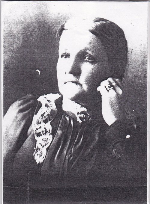 A woman in a dark high-necked dress with a lace collar, one hand raised to her cheek, rings on her fingers, photographed in a studio portrait.