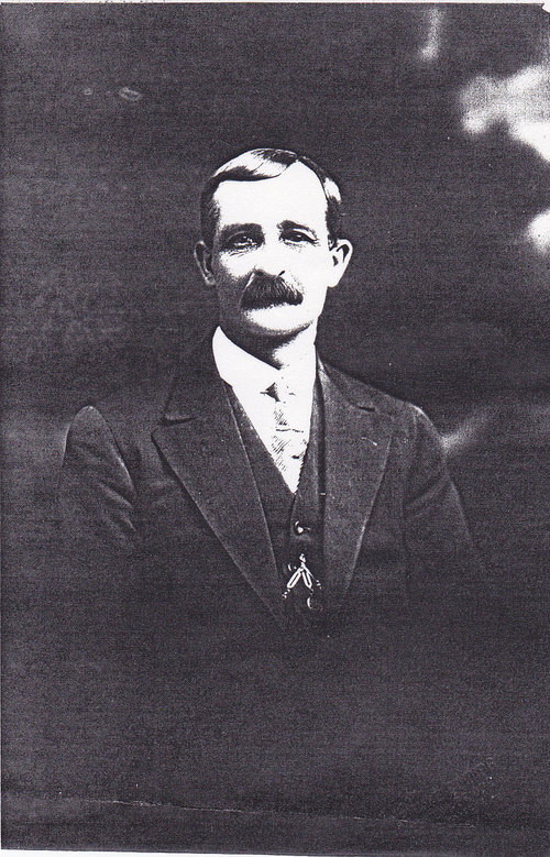 A moustached man in a dark suit and tie, with a watch chain at his waistcoat, photographed side-on in a studio portrait.