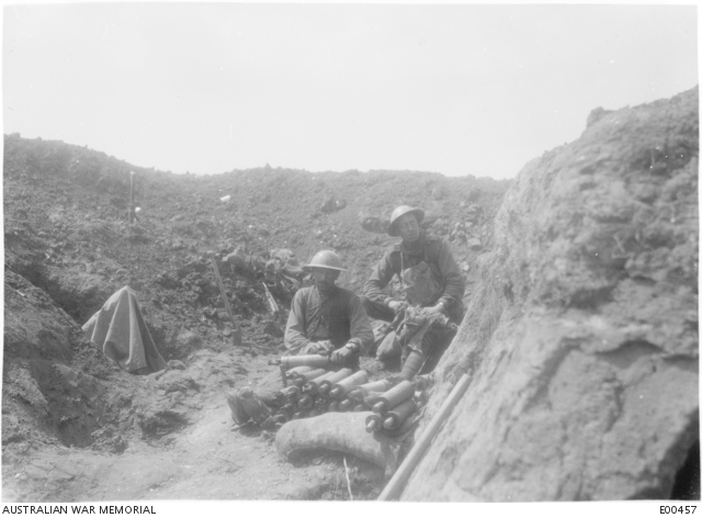 Two Australian soldiers in steel helmets seated in a shell hole beside a stacked pile of artillery and mortar shells.