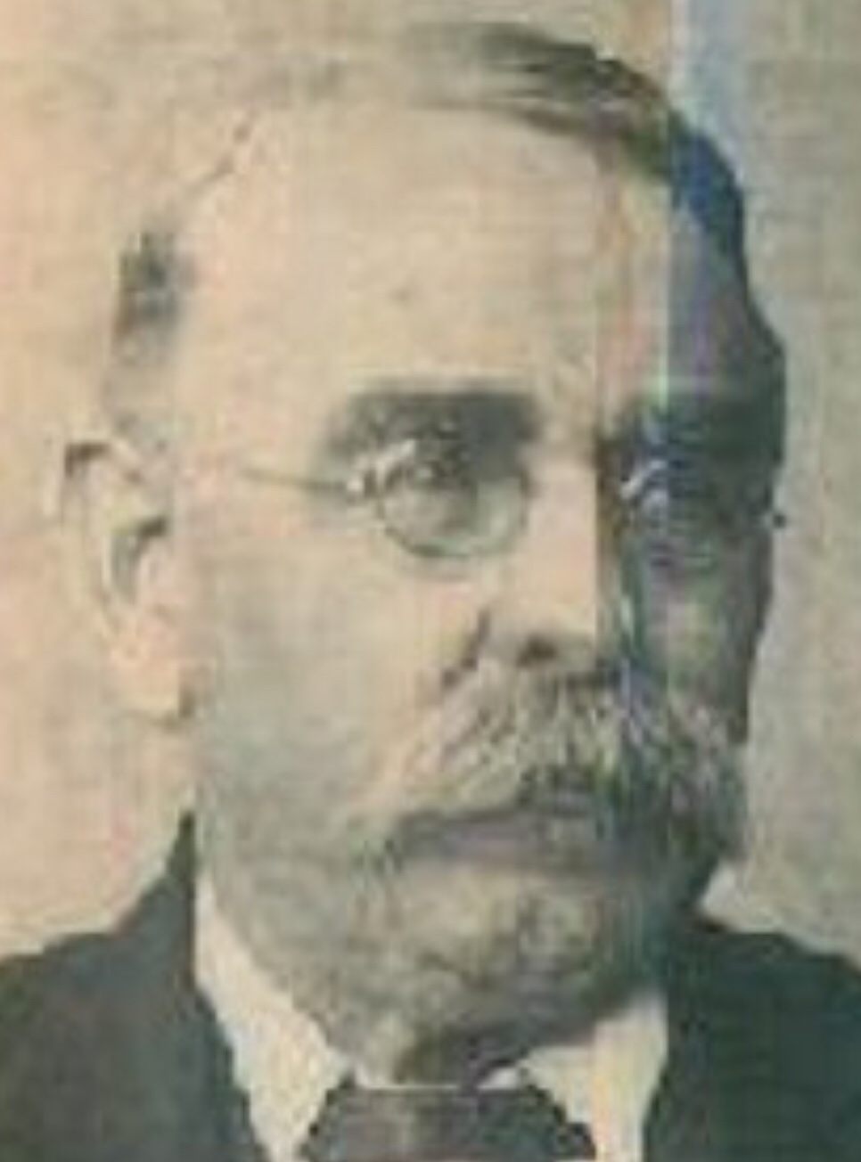 A balding, bespectacled man with a moustache and short beard, in a dark suit and high collar, in an old studio portrait.
