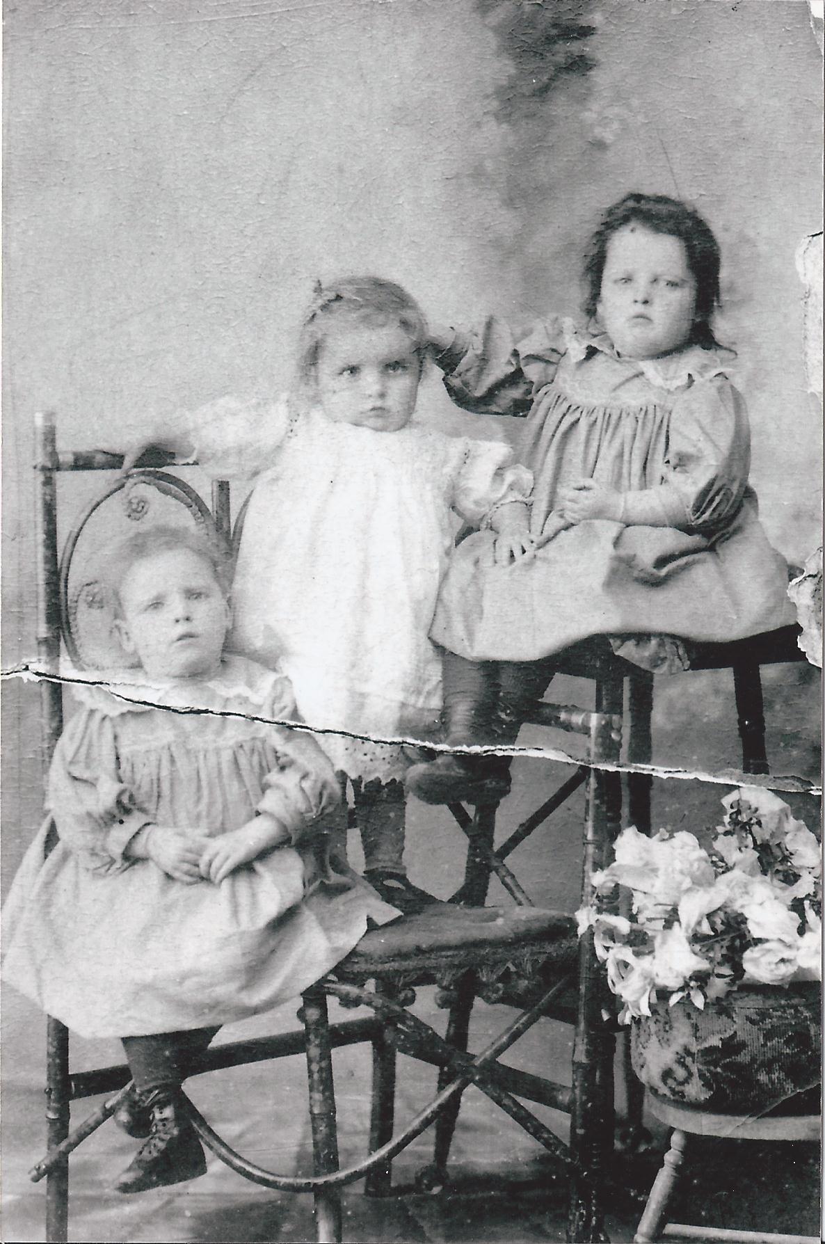 A torn, damaged studio photograph of three small children in white Edwardian dresses, two standing on ornate bamboo chairs and one seated, with a vase of flowers at the foot of the picture.