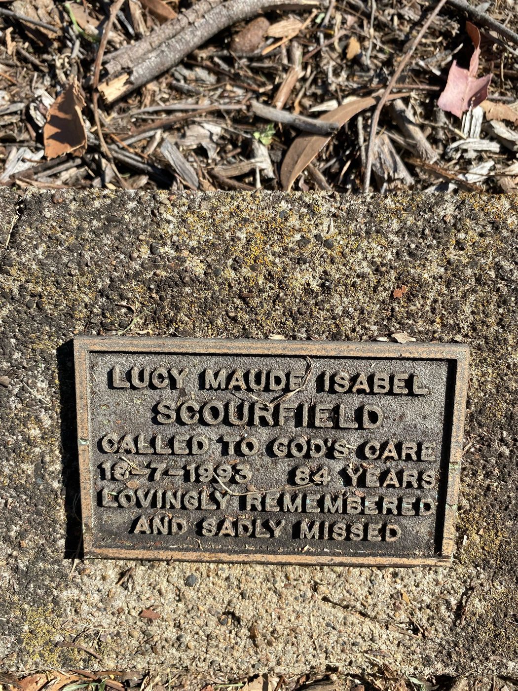 A small bronze memorial plaque set in a garden bed, inscribed to Lucy Maude Isabel Scourfield, died 18 July 1993.