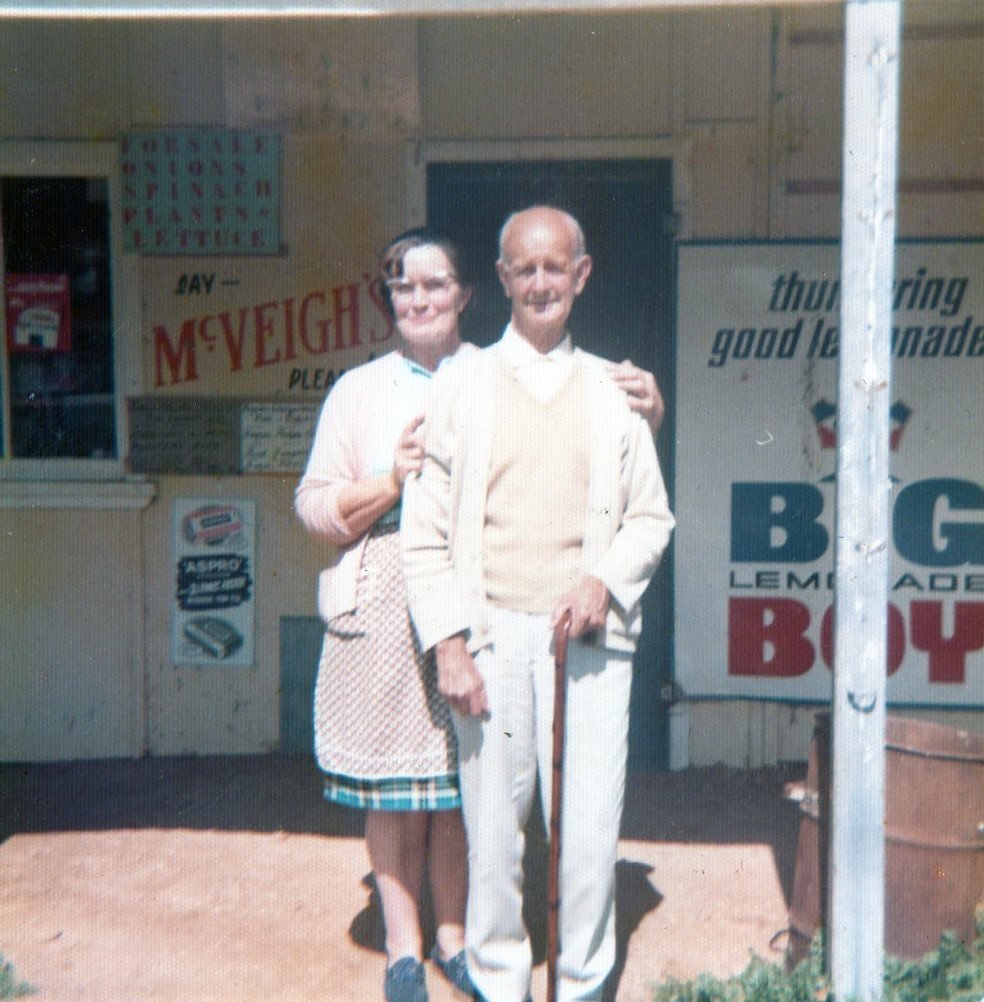 A middle-aged woman with her arm around the shoulders of an elderly man leaning on a walking cane, standing together outside a country general store.
