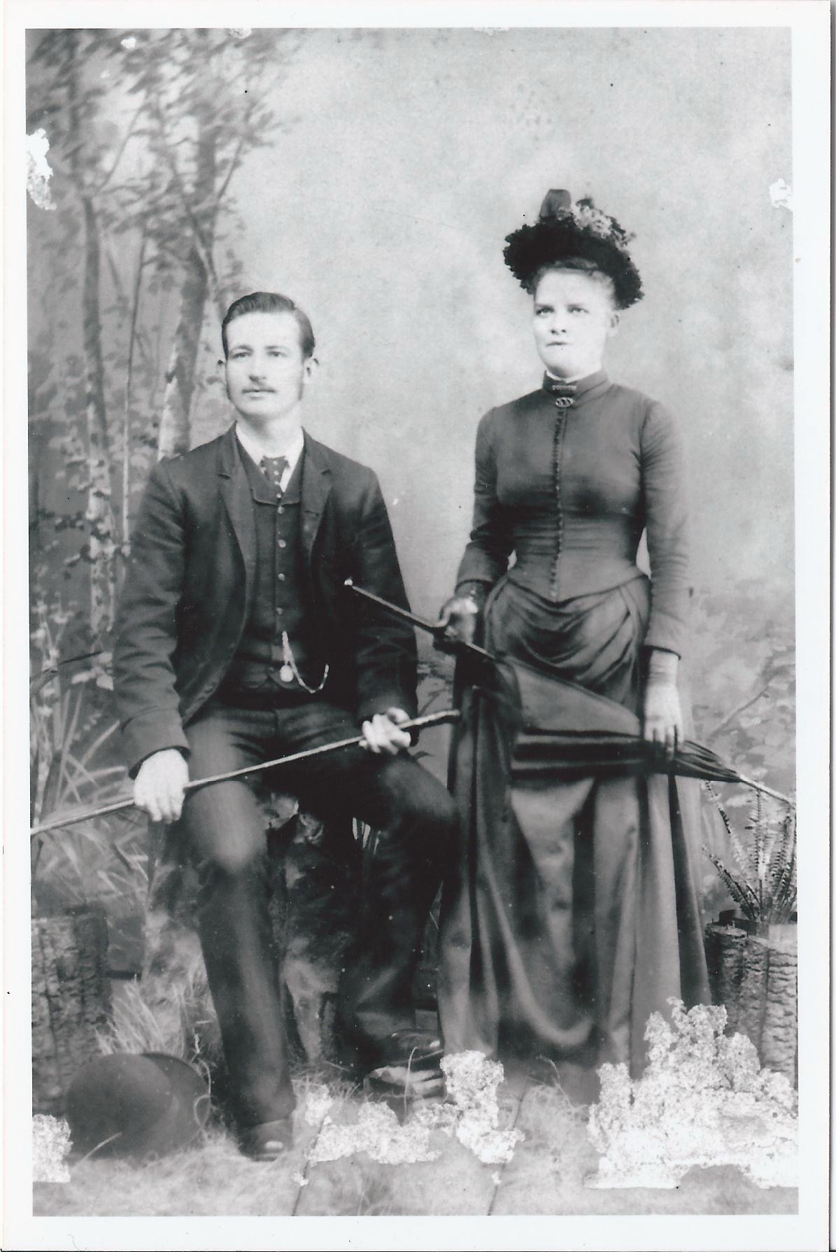 A young man seated and a young woman standing beside him, in Victorian dress, he holding a cane and she a furled umbrella, photographed in a studio against a painted backdrop.