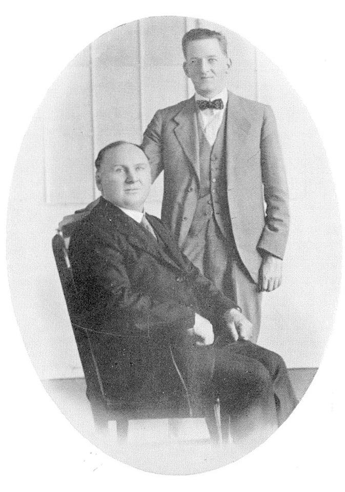 Oval studio photograph of a heavy-set older man seated in a wooden chair and a younger man in a light three-piece suit and bow tie standing beside him with a hand on the chair back.