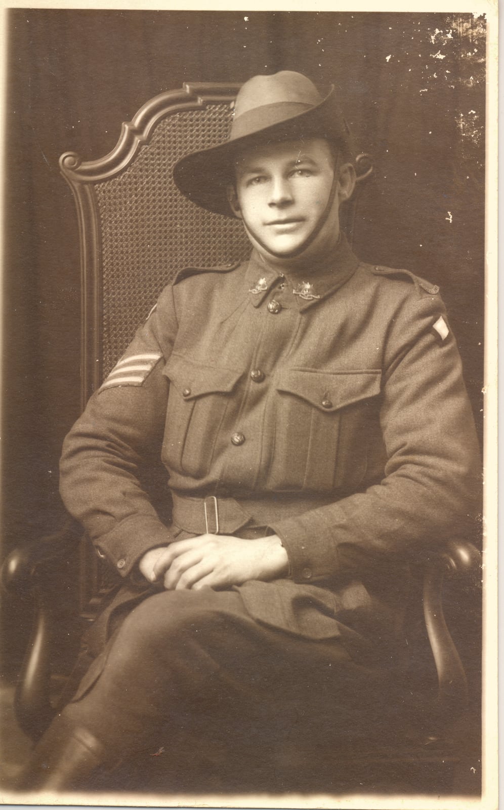 Sepia studio portrait of a young Australian soldier seated in a cane-backed chair, in service dress and slouch hat with chin strap, with sergeant’s chevrons on his right sleeve.