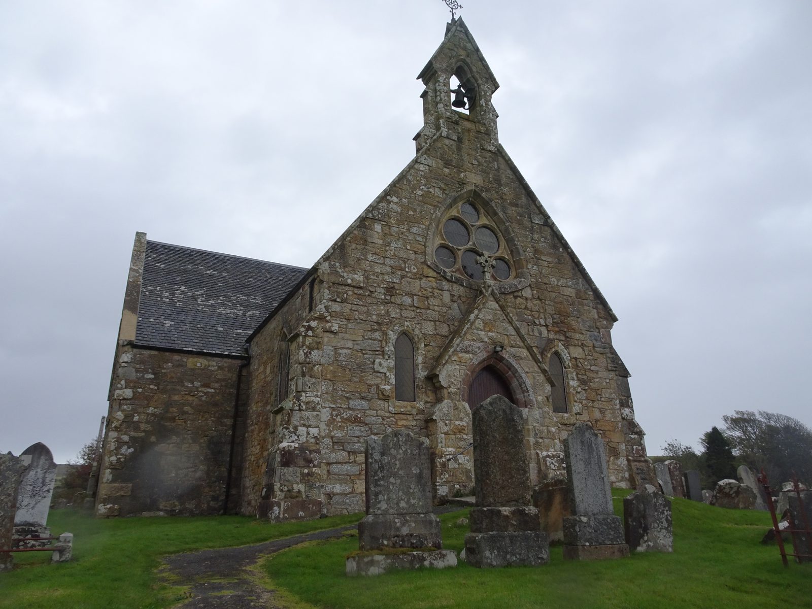A small stone church with a rose window and a bellcote, standing among old gravestones under a stormy sky.