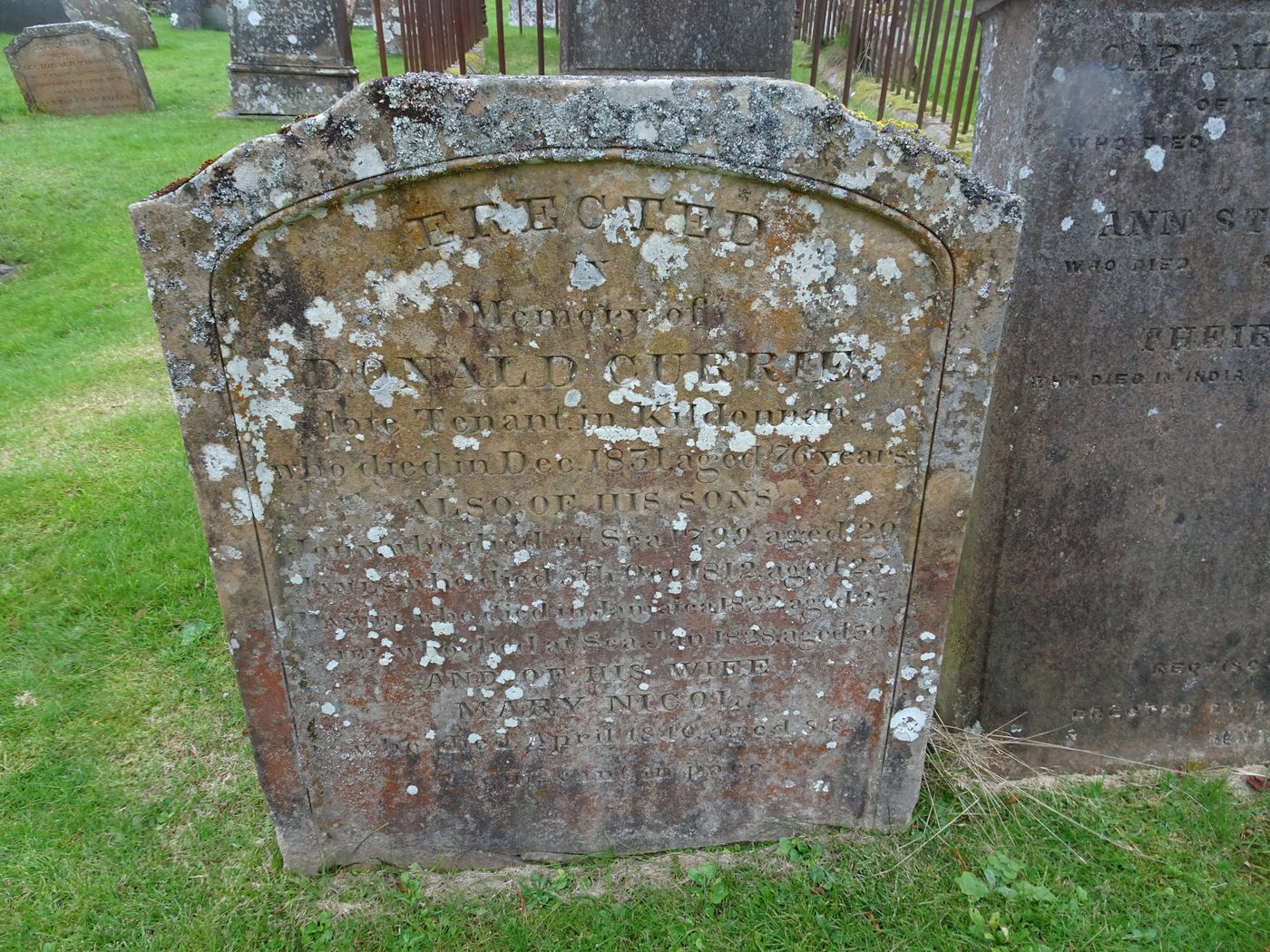 A lichen-spotted sandstone headstone: Erected in memory of Donald Currie, late tenant in Kildonan, who died in December 1831 aged 76 years, also of his sons, two of whom died at sea and one in Jamaica, and of his wife Mary Nicol.