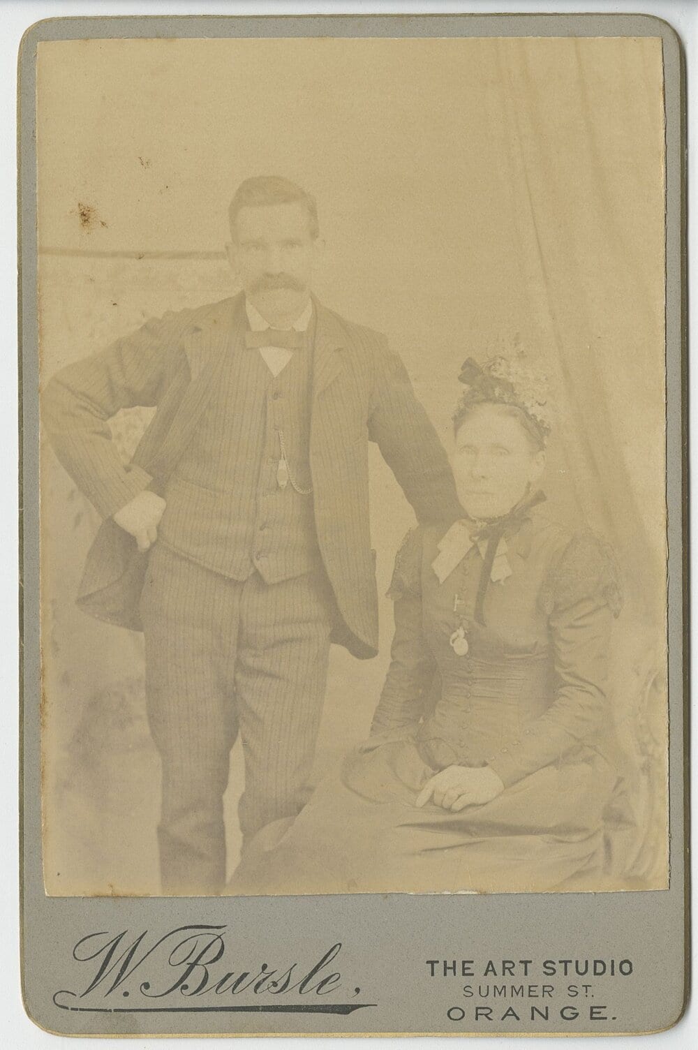 Cabinet-card studio portrait of a moustached man standing with one hand on his hip beside a seated older woman in a dark high-necked Victorian dress and a flowered bonnet.