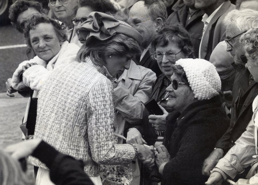 Black and white press photograph of a crowd at a barrier; a woman in a light tweed suit shakes hands with an older woman in a white crocheted hat and dark glasses.