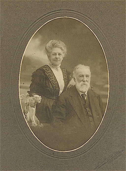 Oval studio portrait on a grey mount: a seated white-bearded man and, standing behind him, a woman in spectacles and a dark braided bodice, a vase of flowers beside them.
