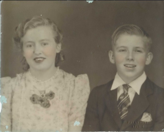Hand-tinted studio photograph of a girl in a floral dress with a ribbon bow and a boy in a suit and striped tie, both smiling.