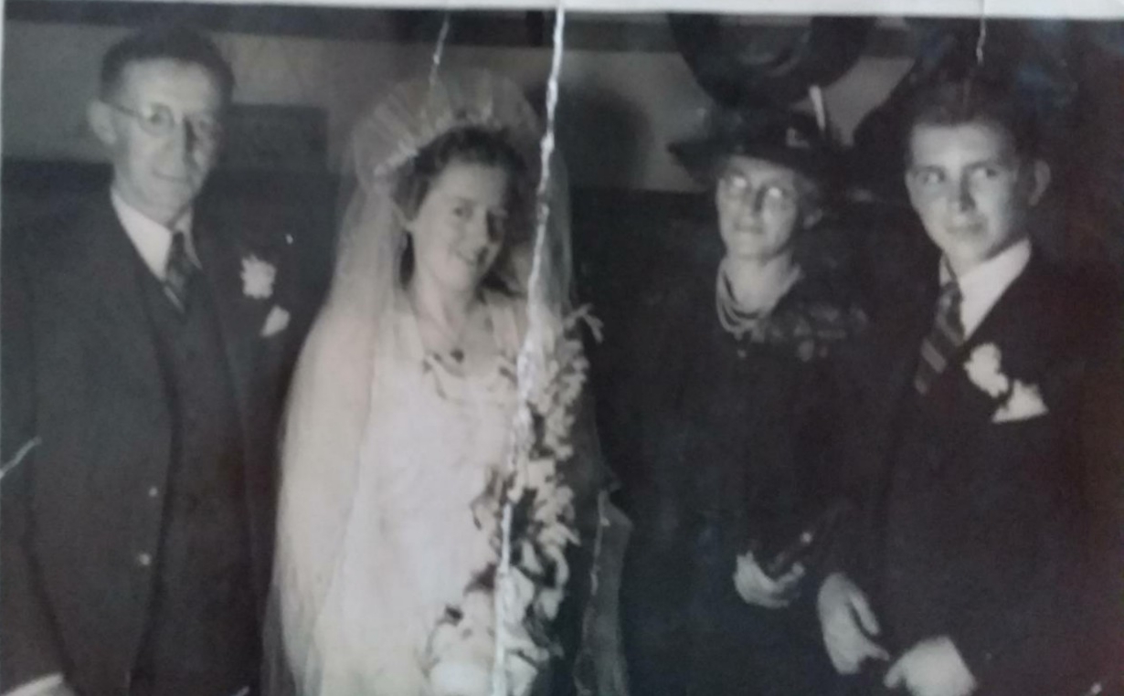 Group photograph of four: a bespectacled man in a suit, a bride in a veil holding a bouquet, a woman in a dark coat and hat wearing spectacles, and a young man in a suit with a buttonhole.