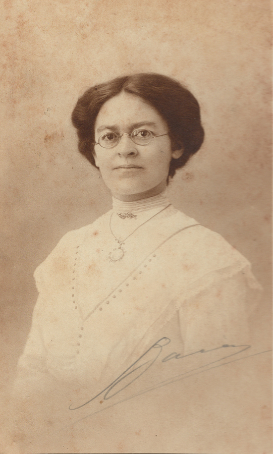 Sepia postcard portrait of a woman in wire-rimmed spectacles and a pale high-necked blouse, signed across the lower right in ink.