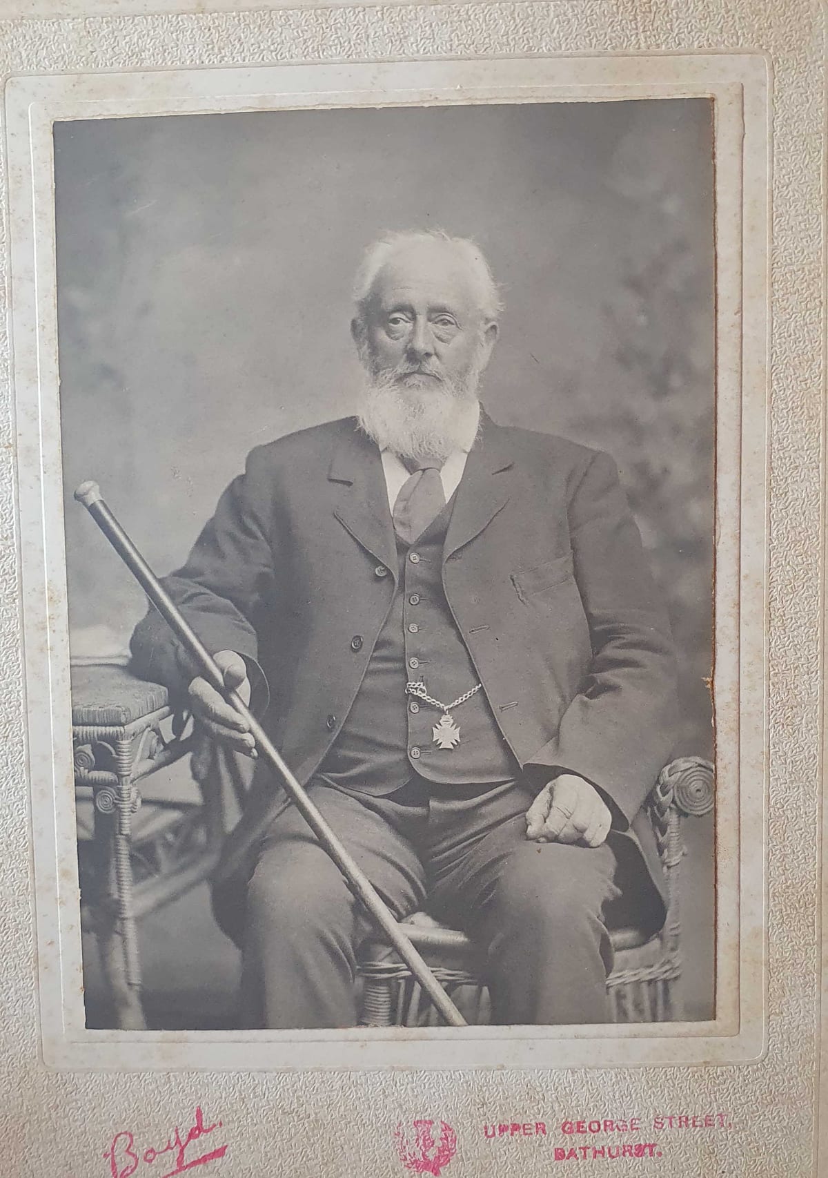Studio cabinet-card portrait of a white-bearded elderly man in a three-piece suit, seated in a cane chair with a walking cane across his lap and a cross-shaped medal hanging from his watch-chain.