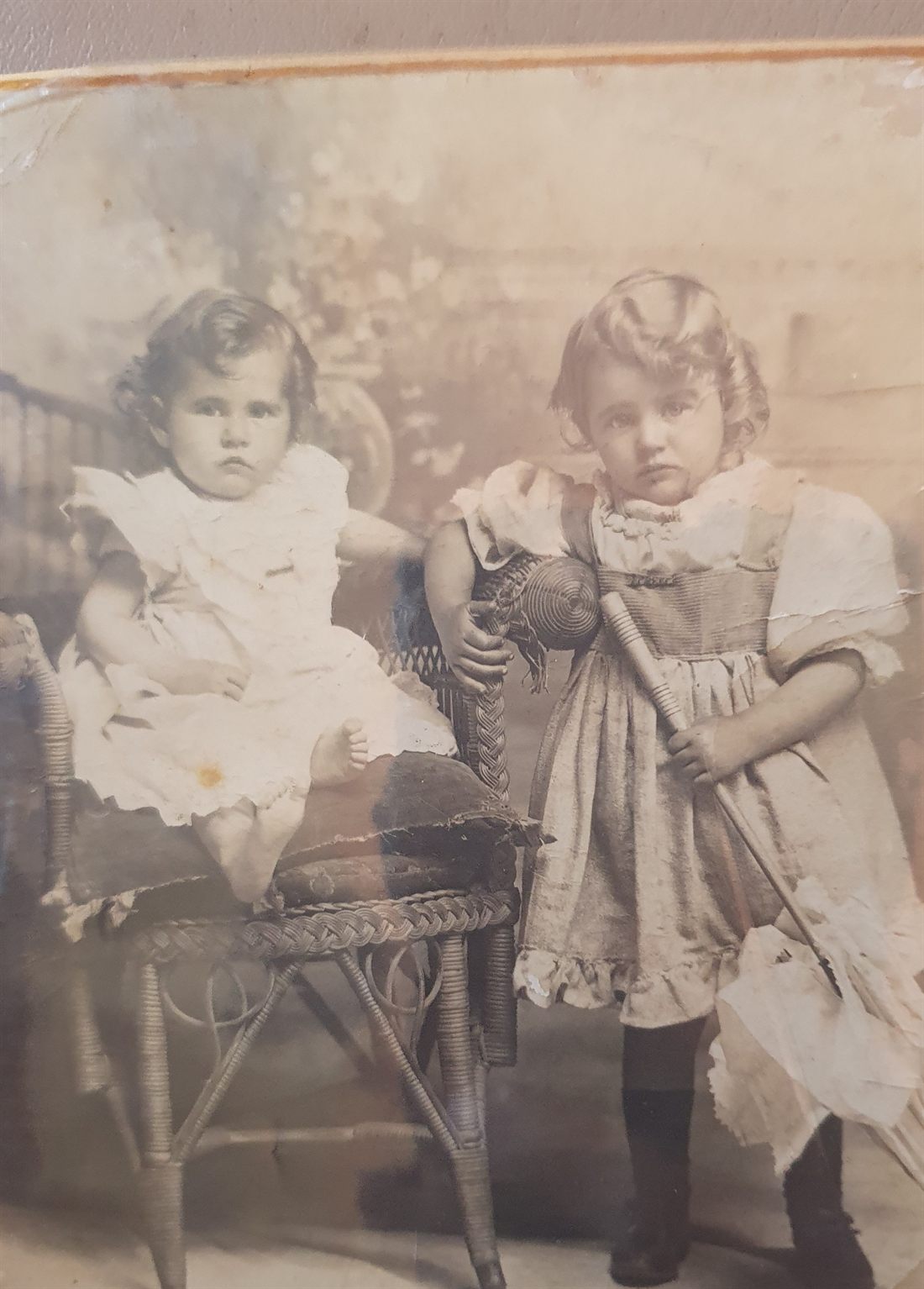 Studio photograph of two small girls: a toddler in a white dress seated on a wicker chair, and an older girl of about four standing beside her in a pinafore, holding a parasol.