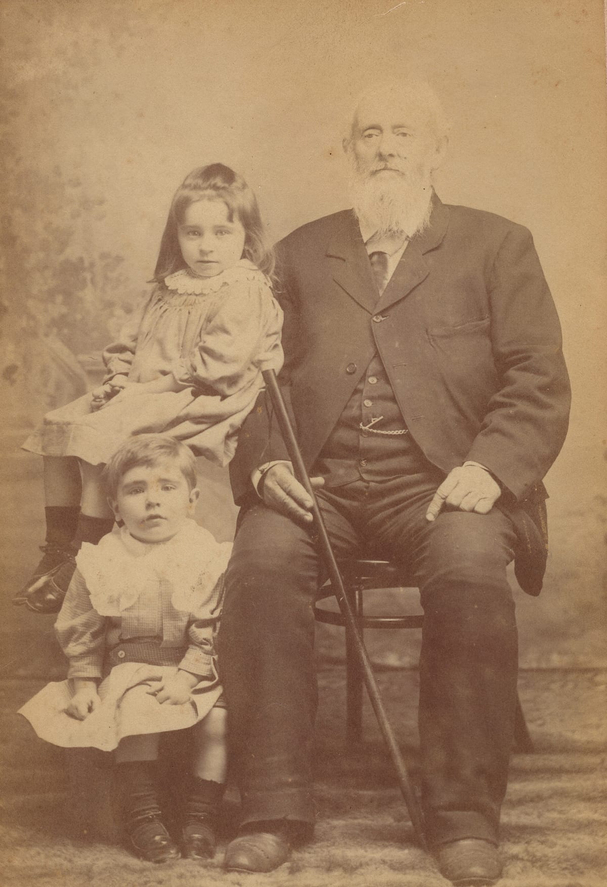 Cabinet-card photograph of a white-bearded elderly man seated with a cane, a small girl in a pale dress perched on the chair-arm beside him and a younger child in white standing at his knee.