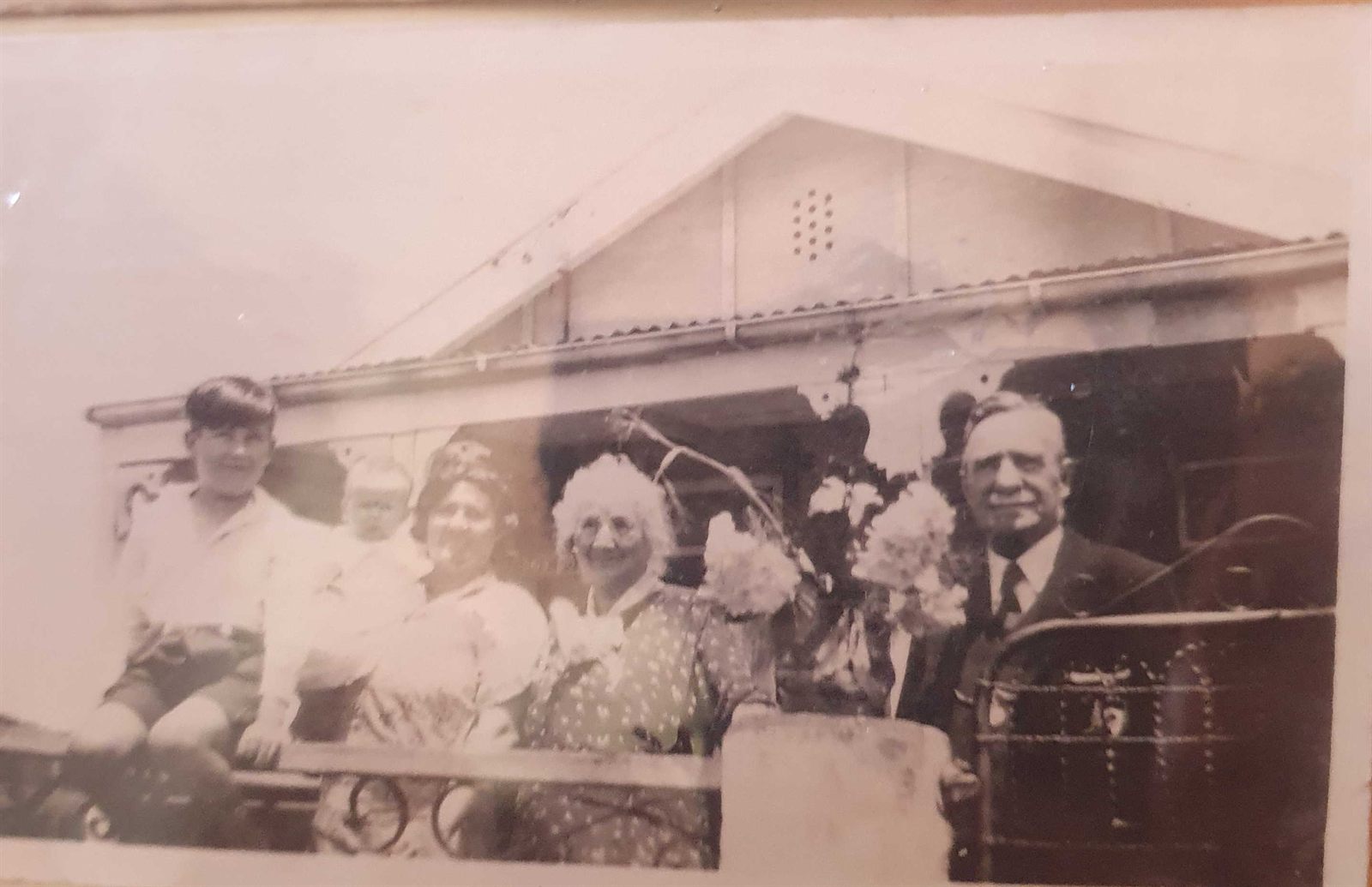 Snapshot of five people and a motor car outside a weatherboard house: a boy in shorts, a woman holding a baby, a second woman, a white-haired elderly woman, and, at the wheel, a bald elderly man in a dark suit and tie.
