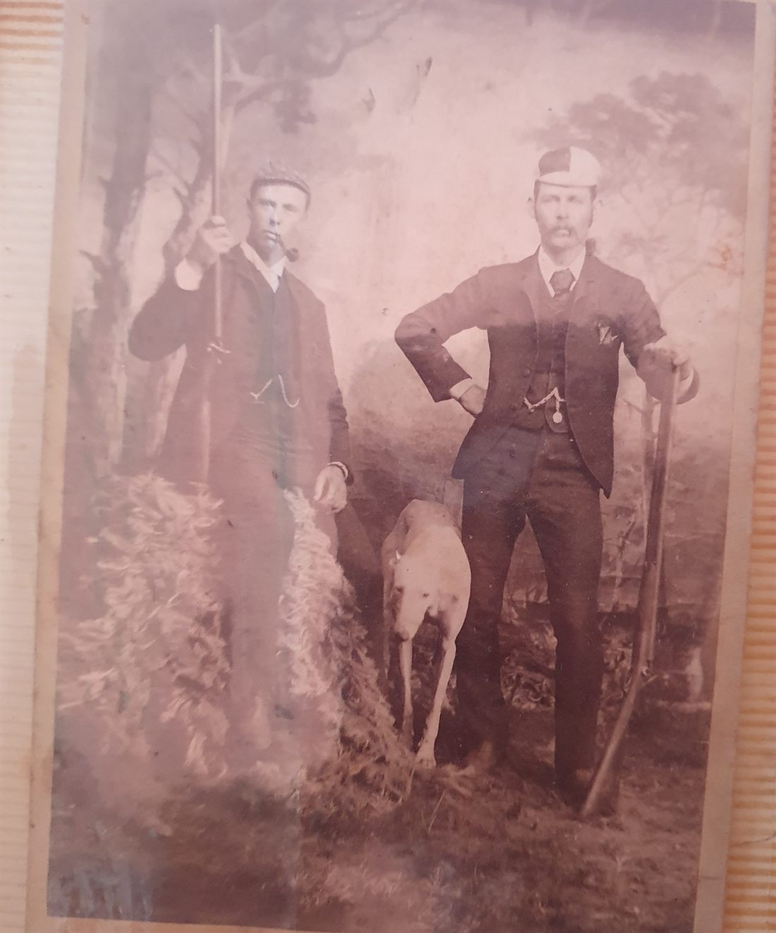 Sepia cabinet-card photograph of two young men in suits and caps posed against a painted woodland backdrop, each holding a long sporting gun, with a whippet-like dog standing between them; the man at right rests one hand on his hip.