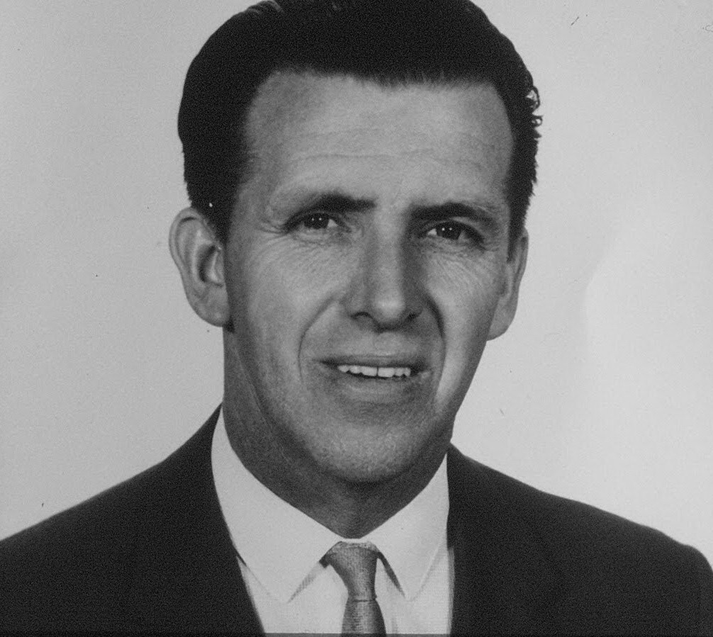 Studio head-and-shoulders portrait of a dark-haired man in a suit and patterned tie, smiling slightly.