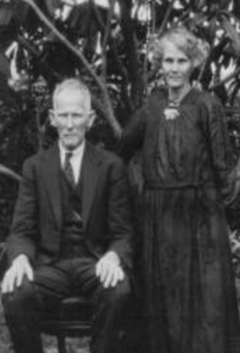 An elderly white-haired man in glasses and a three-piece suit, seated, with a grey-haired woman in a long dark dress standing beside him in a garden.