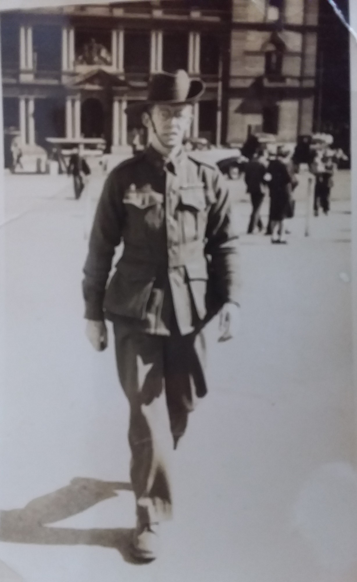 Photograph of a bespectacled man in slouch hat and service dress walking towards the camera across a paved forecourt, a large classical stone building behind him and passers-by in coats and hats.