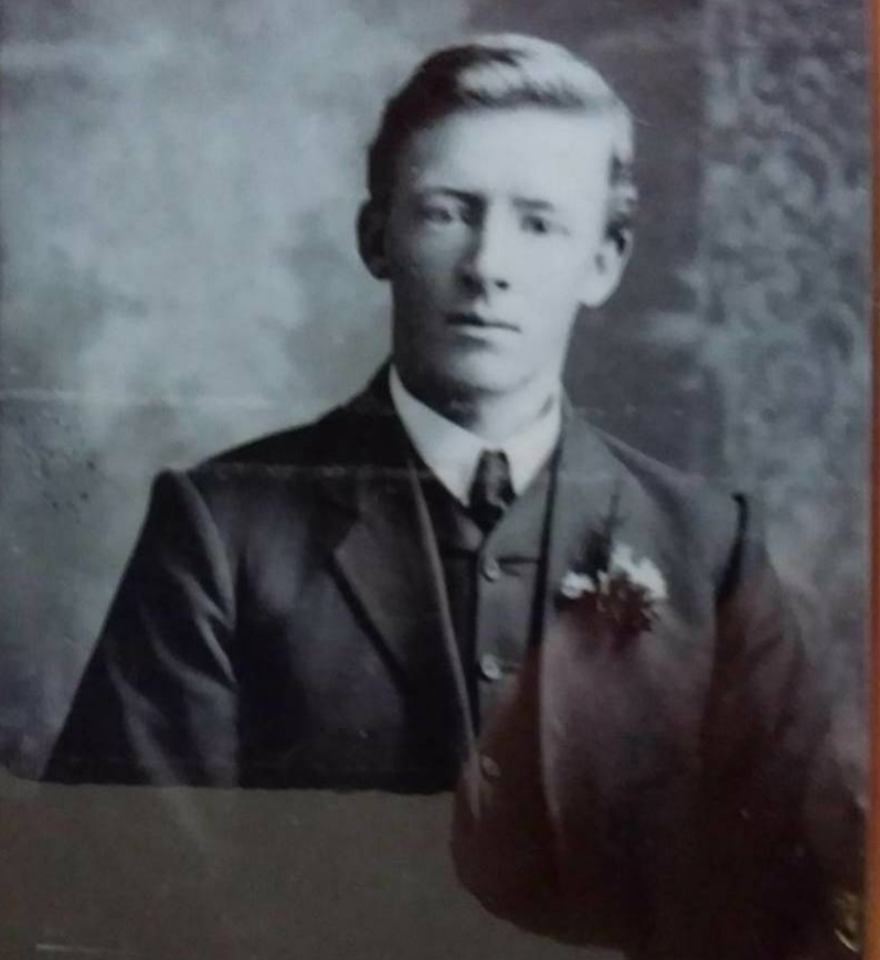 Sepia studio half-length portrait of a young man in a dark three-piece suit and tie, with a light flower in his buttonhole.
