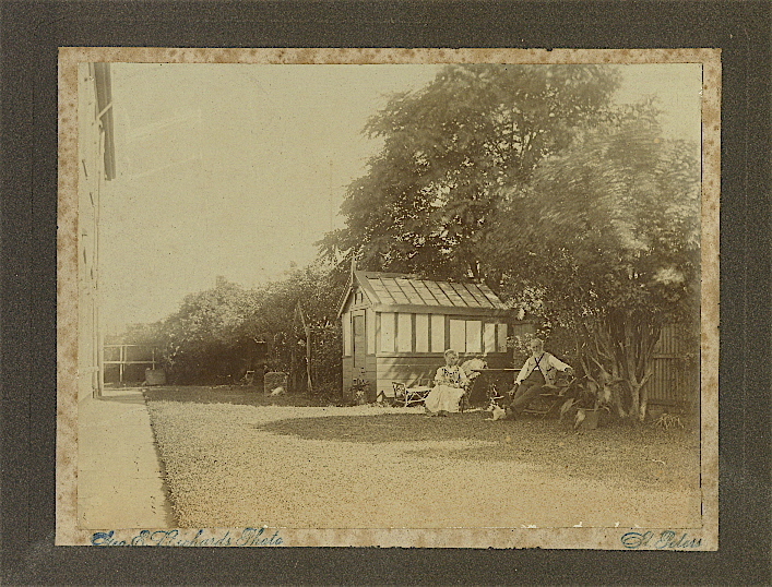 Faded albumen photograph on a card mount: a gravel drive beside a house, a large tree, and an elderly couple seated by a small timber-and-glass garden house.
