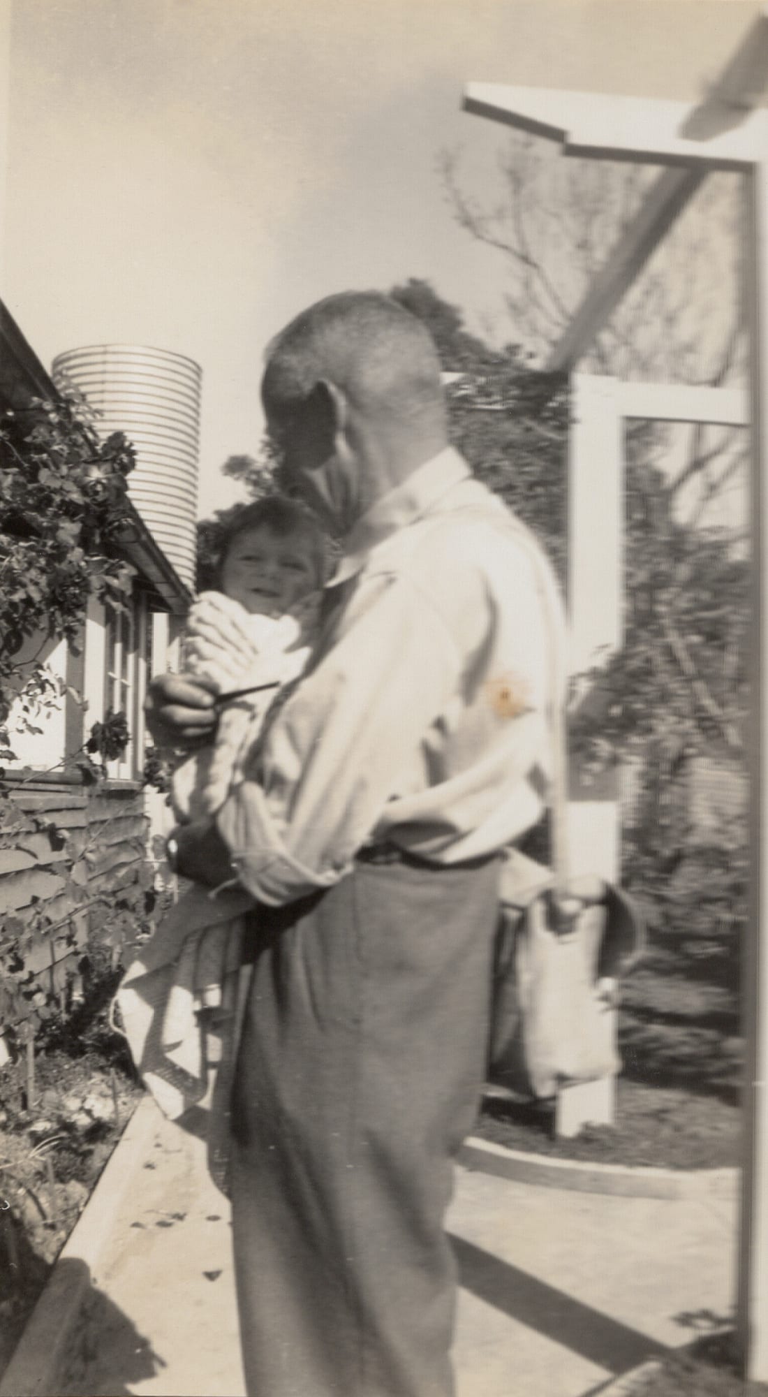 A man in shirtsleeves and work trousers, a bag slung on a post behind him, stands holding a small baby against his shoulder beside a weatherboard house with a corrugated-iron rainwater tank and a grapevine on wires.