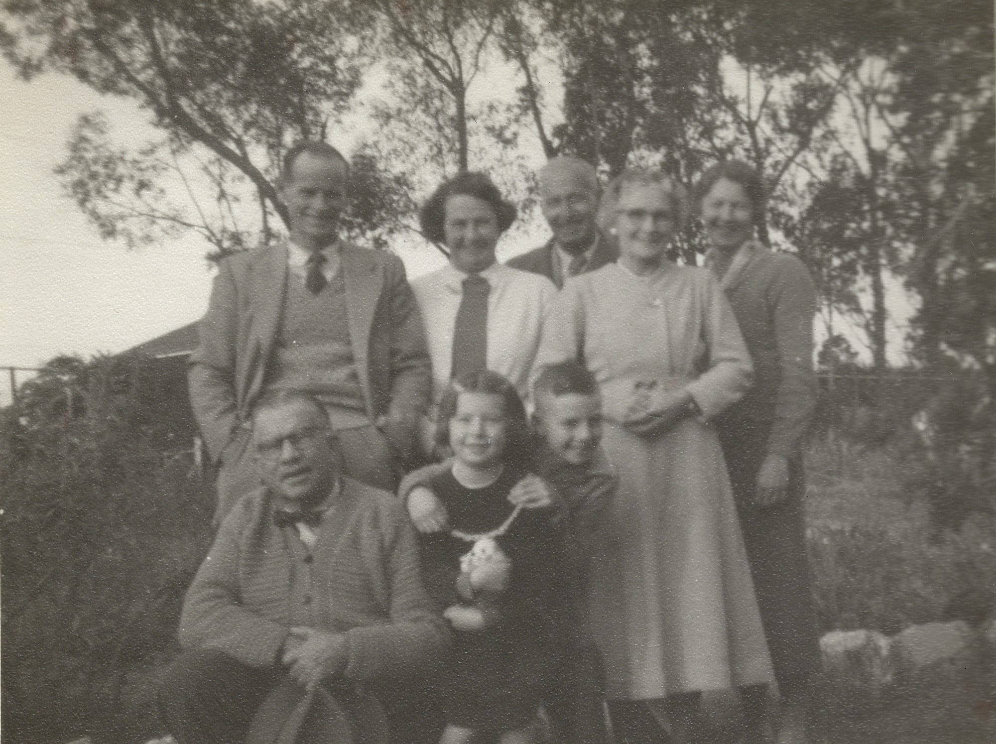 A group of adults outdoors on a hillside with trees behind — five standing, an older bespectacled man seated at left, and two children in front.