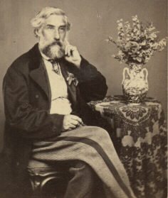 Carte-de-visite of a bearded man in a dark coat, seated beside a table with a vase of flowers, his hand raised to his chin.