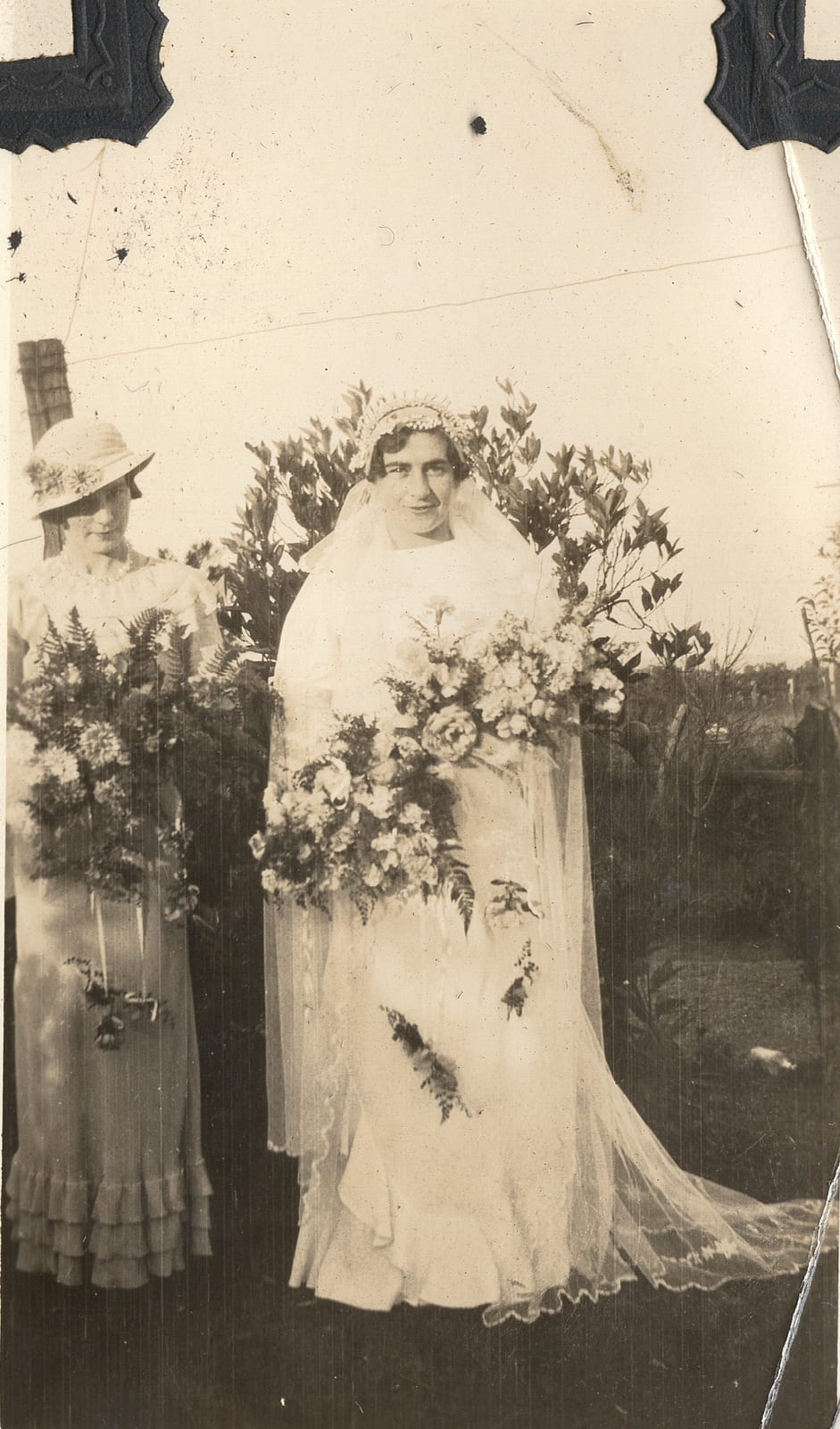 Outdoor photograph of a bride in a full-length gown and long veil holding a large cascade bouquet, beside a young woman in a light tiered dress and a soft hat holding a fern-and-flower posy, garden shrubbery behind.