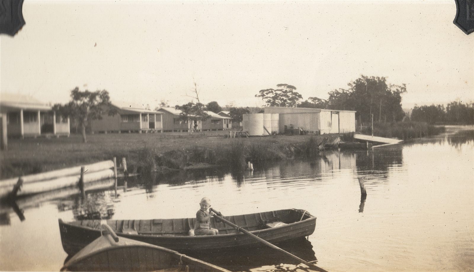 A calm lake with a row of identical timber cabins raised on stumps along the far bank, water tanks and bush behind; in the foreground a small boy sits in a wooden dinghy holding an oar.