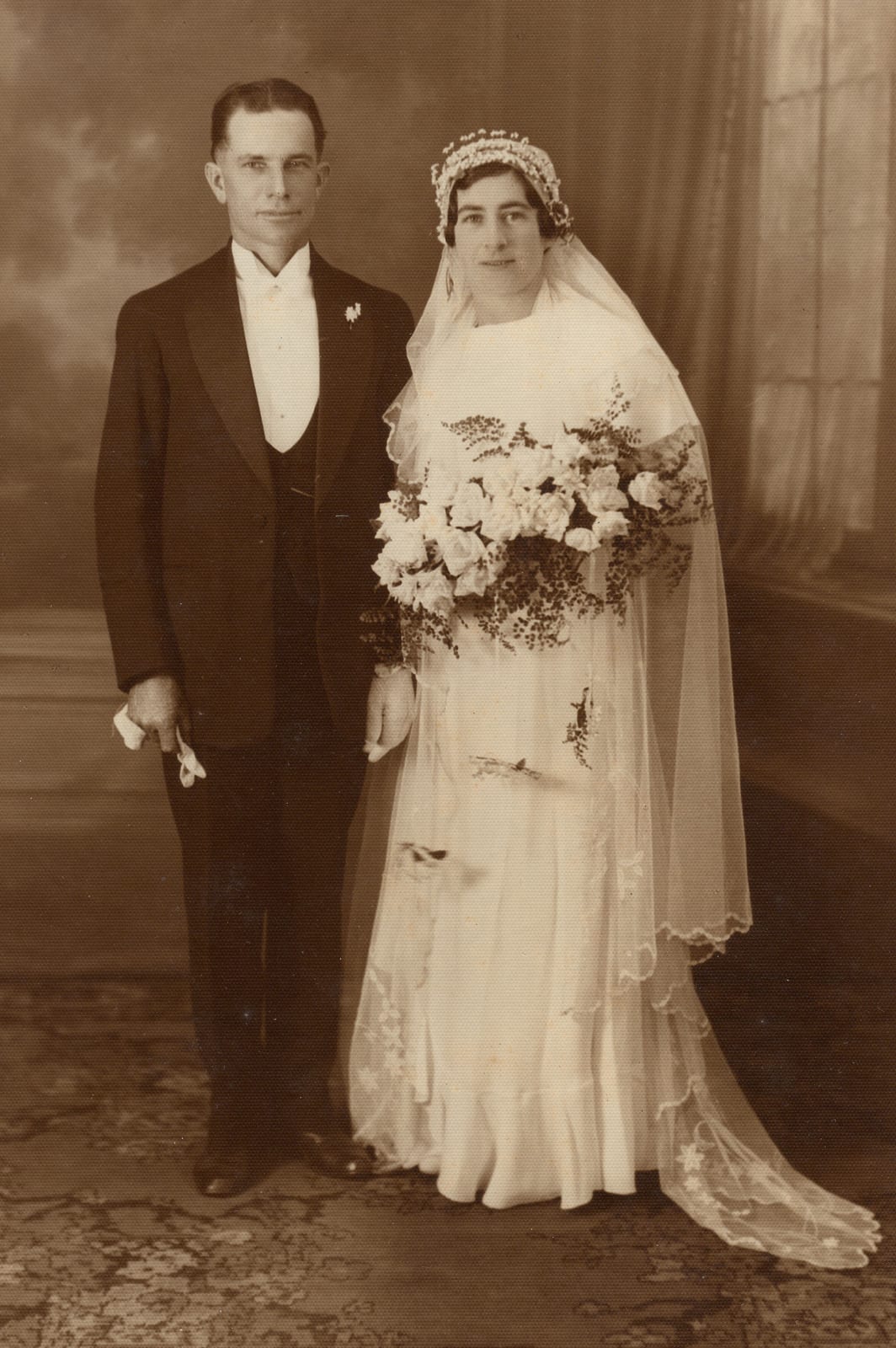 Studio wedding portrait: a fair-haired man in a dark three-piece morning suit with a white bow tie and a sprig of wattle at the lapel, standing beside a bride in a bias-cut satin gown under a long lace-edged veil, holding a cascade bouquet of flowers and maidenhair fern.
