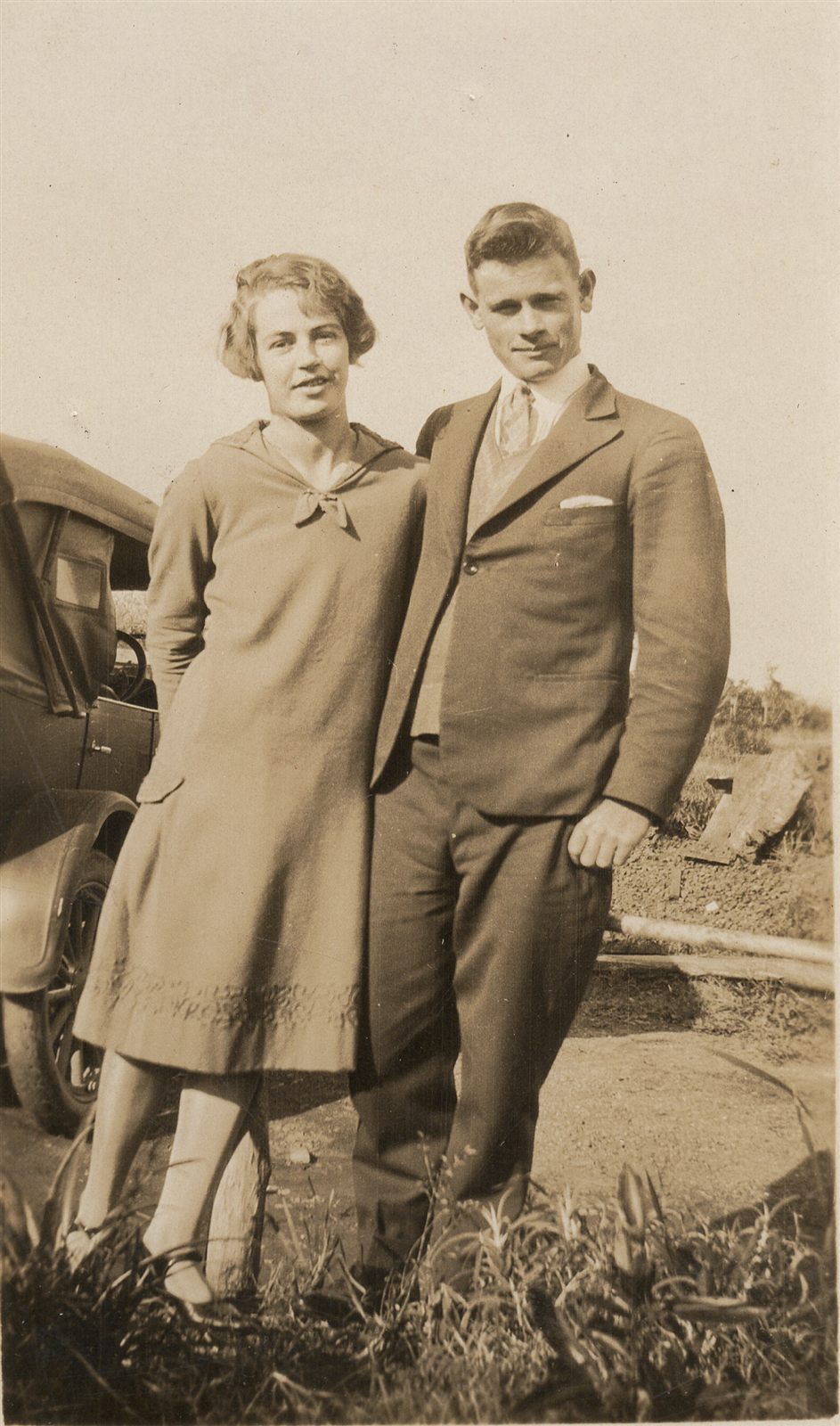 A young woman in a drop-waisted dress with a sailor collar stands beside a young man in a three-piece suit, an open touring car behind them and paddock beyond.