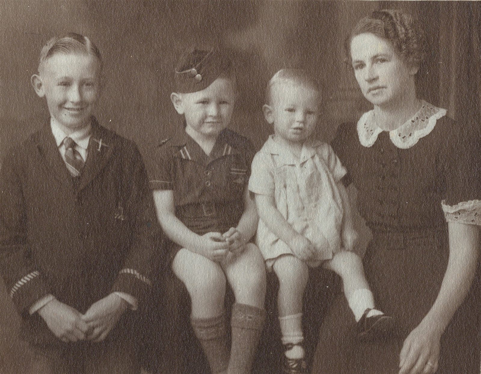 A studio portrait of a woman seated with three boys — an older boy in a school tie, a middle boy in a soft forage cap, and a toddler in a romper on the middle boy’s knee.