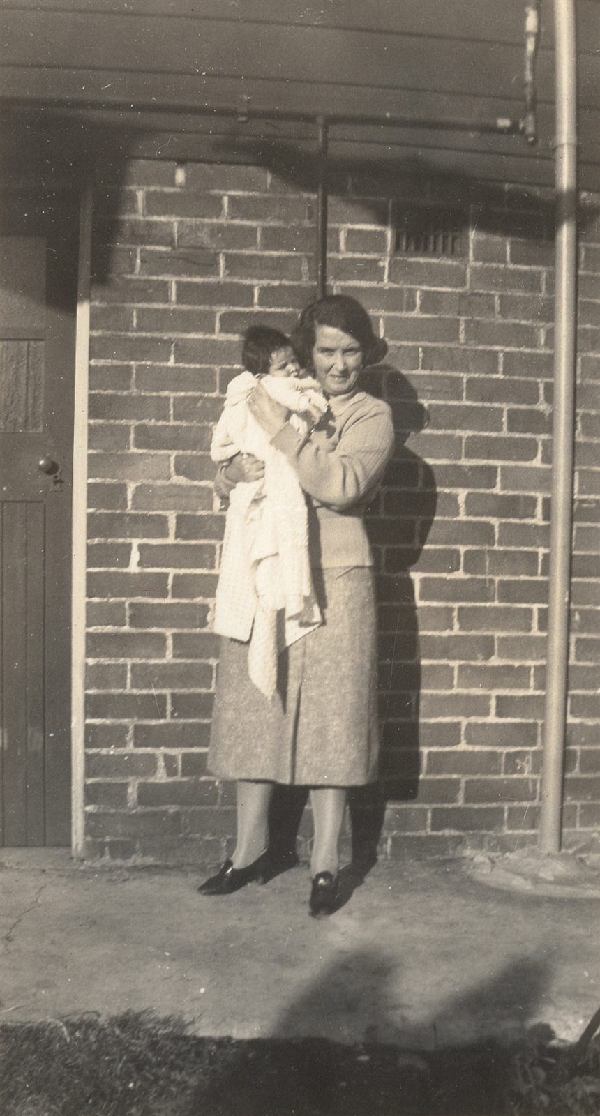 A woman in a knitted jumper and skirt stands against a brick wall holding a small baby wrapped in a shawl.