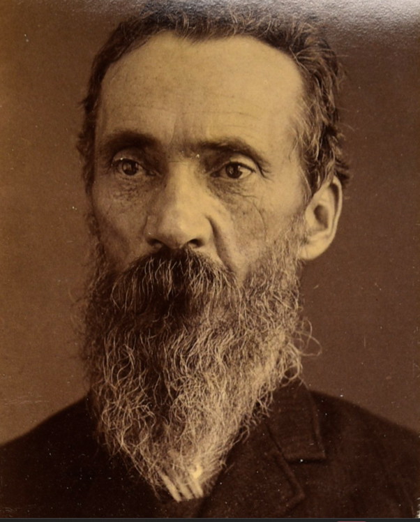 A formal, heavily bearded Victorian-era studio-style photograph of an older man with brown hair, grey eyes and a long grey-flecked beard, wearing a dark coat — cropped from a NSW gaol description sheet.