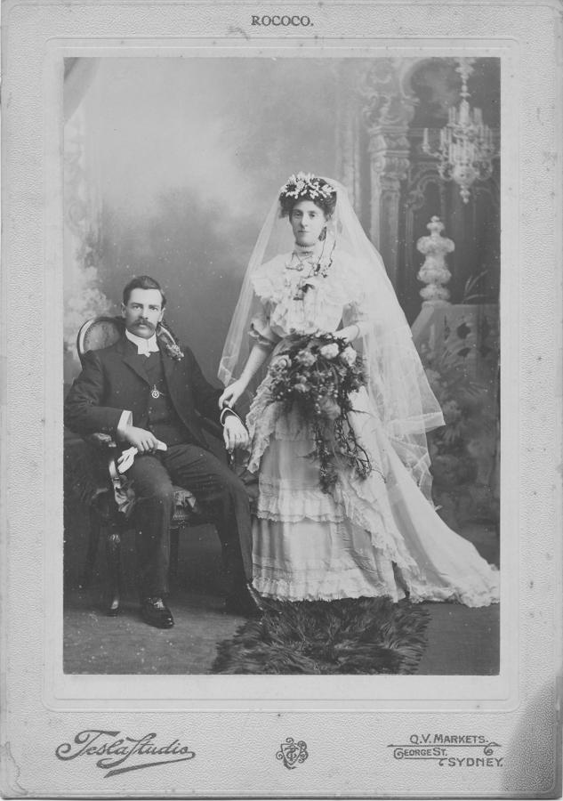 Studio wedding photograph on a printed mount: a seated moustached man in a dark suit and a standing bride in a tiered white gown with a long veil and a trailing bouquet.